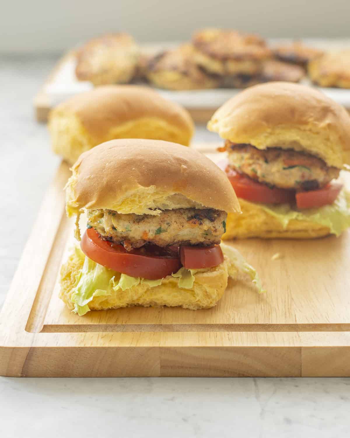 Three slider buns on wooden chopping board wth chicken patty, tomato and lettuce inside. Chicken patties on tray in background.