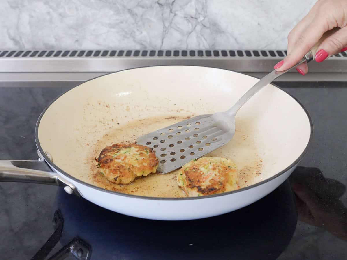 Hand holding a metal spatula flipping chicken patty over in a pan.