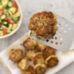 Fried chicken patty held up on a metal spatula, a dozen patties below on wooden chopping board, fresh salad in bowl on benchtop.