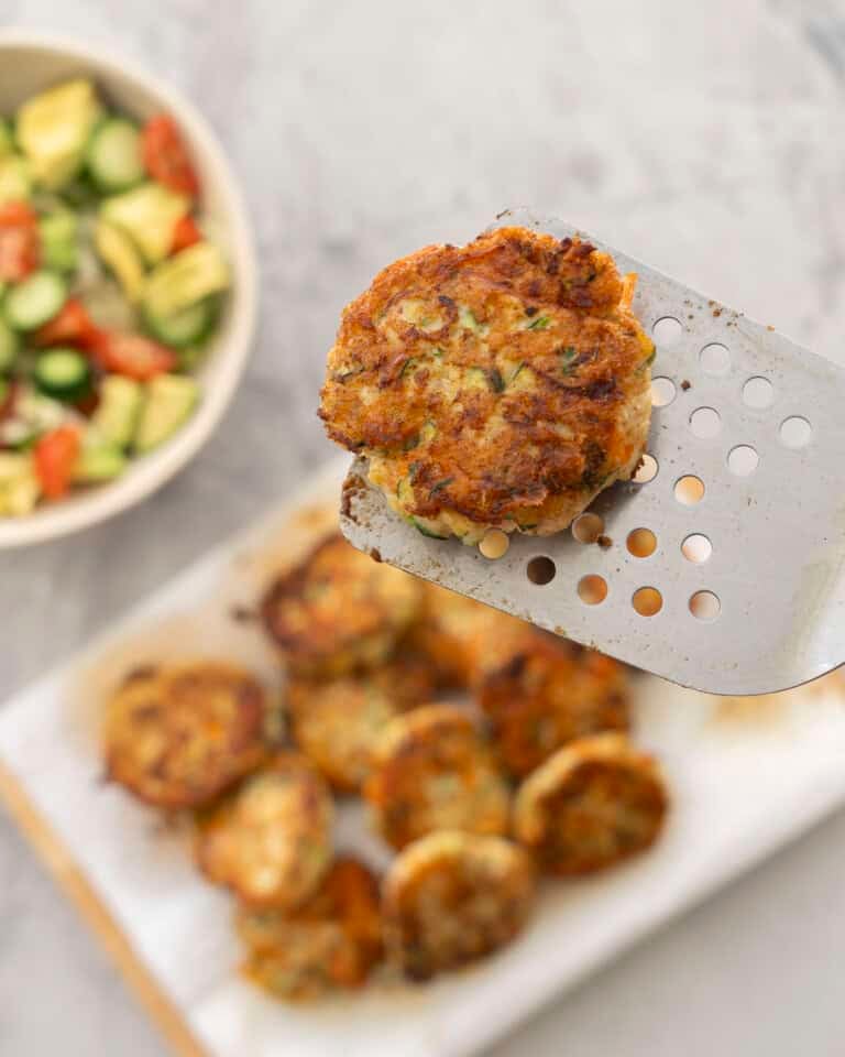 Fried chicken patty held up on a metal spatula, a dozen patties below on wooden chopping board, fresh salad in bowl on benchtop.