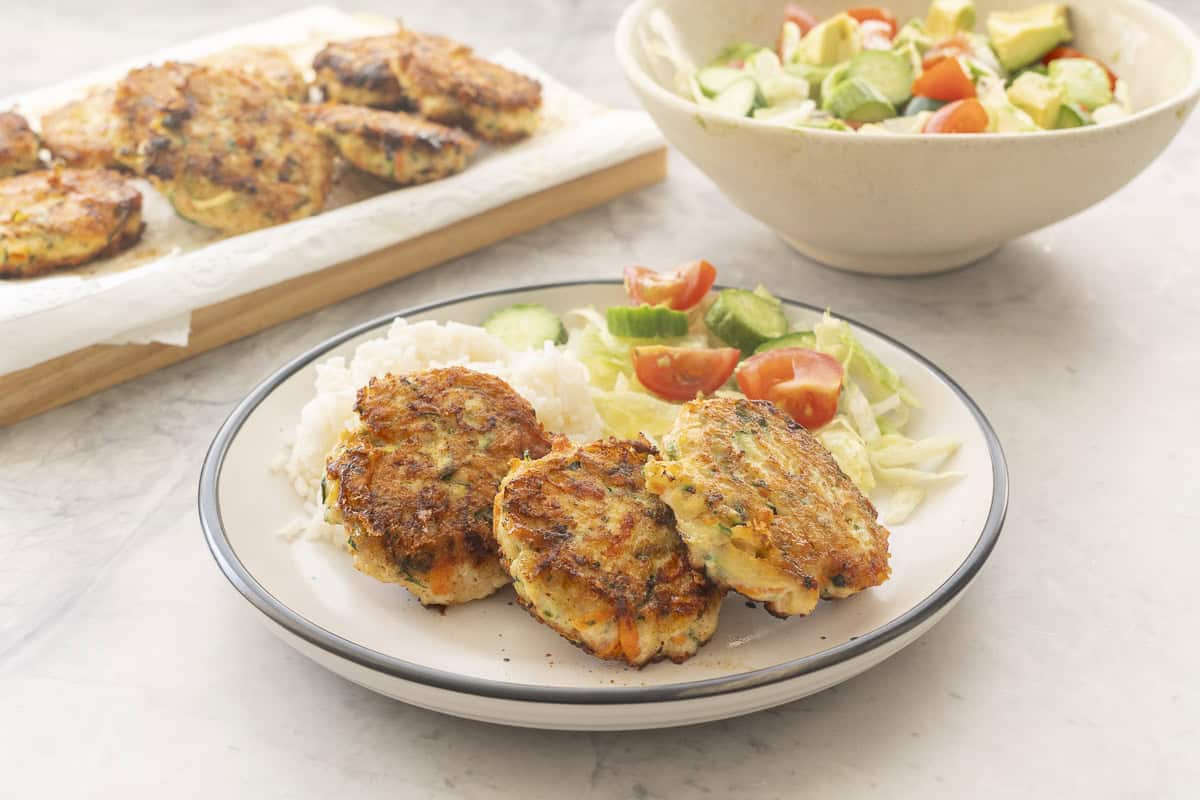 Three chicken patties, a fresh salad and small portion of rice on a dinner plate, Bowl of salad and tray of chicken patties in background.