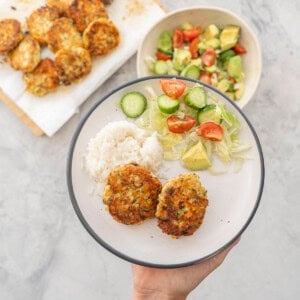 Three chicken patties, a fresh salad and small portion of rice on a dinner plate being held up to camera, Bowl of salad and tray of chicken patties in background on benchtop.