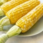 Four cooked corn on the cob on a plate with melted butter and salt and pepper on top.