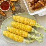 Four cooked corn on the cob on a plate with melted butter and salt and pepper on top, tray of crispy potato wedges with sweet chilli sauce, chicken drumsticks in baking tray.