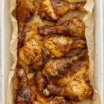 Nine cooked chicken drumsticks with sticky glossy paprika sauce on top inside a white oven dish lined with baking paper.