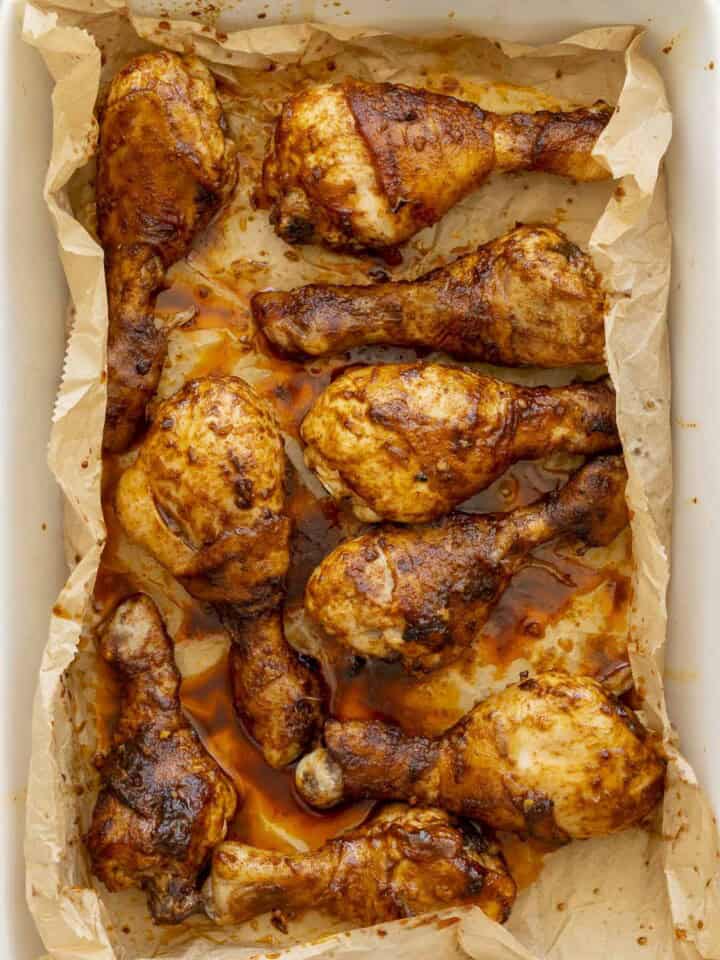 Nine cooked chicken drumsticks with sticky glossy paprika sauce on top inside a white oven dish lined with baking paper.