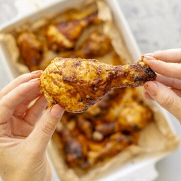 Hand holding up a glossy sticky drumstick above a white baking dish with cooked chicken drumsticks inside.