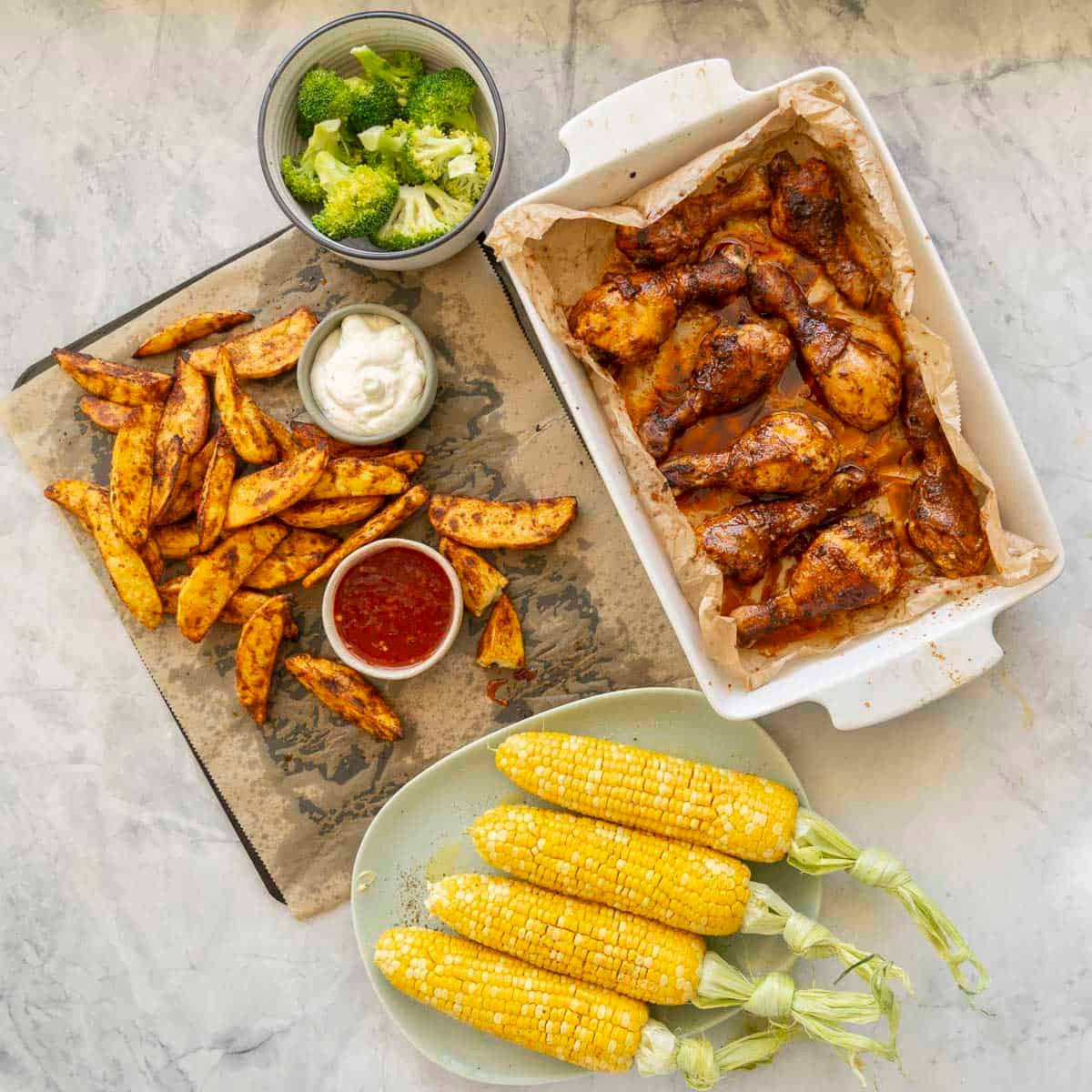 White baking dish with cooked chicken drumsticks inside, roasting tray lined with baking paper with crispy homemade potato chips and two small ramekins with aloli and sweet chilli, a bowl of cooked broccoli florets and four corn on the cob on a plate.