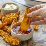 Crispy seasoned potato wedges on lined baking tray with two ramekins, aioli and sweet chilli sauce, hand holding a wedge dipping into sweet chilli sauce.