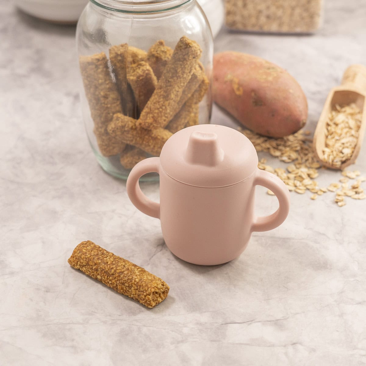 Baby rusk and silicone baby cup, wooden spoon with oats, sweet potato and jar with remainder rusks in background.