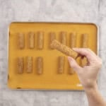 Womans hand holding a cooked baby rusk to camera with rest below on lined baking tray.