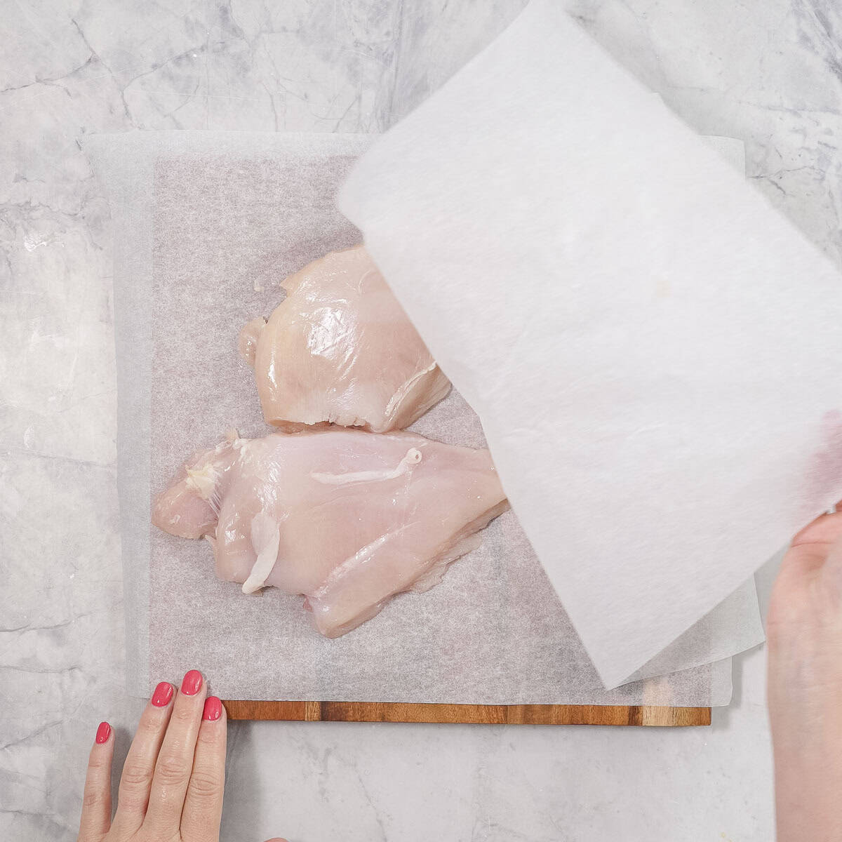 Chicken breasts on wooden chopping board with baking paper, Hand pealing back a piece of baking paper off the top of the breasts.