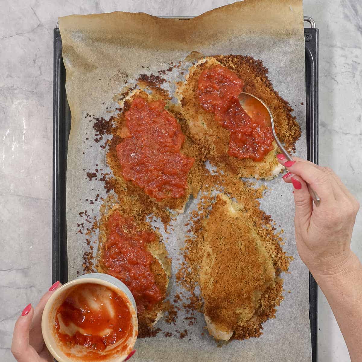 Hand spooning marinara sauce onto crispy parmesan chicken breasts on lined baking tray.