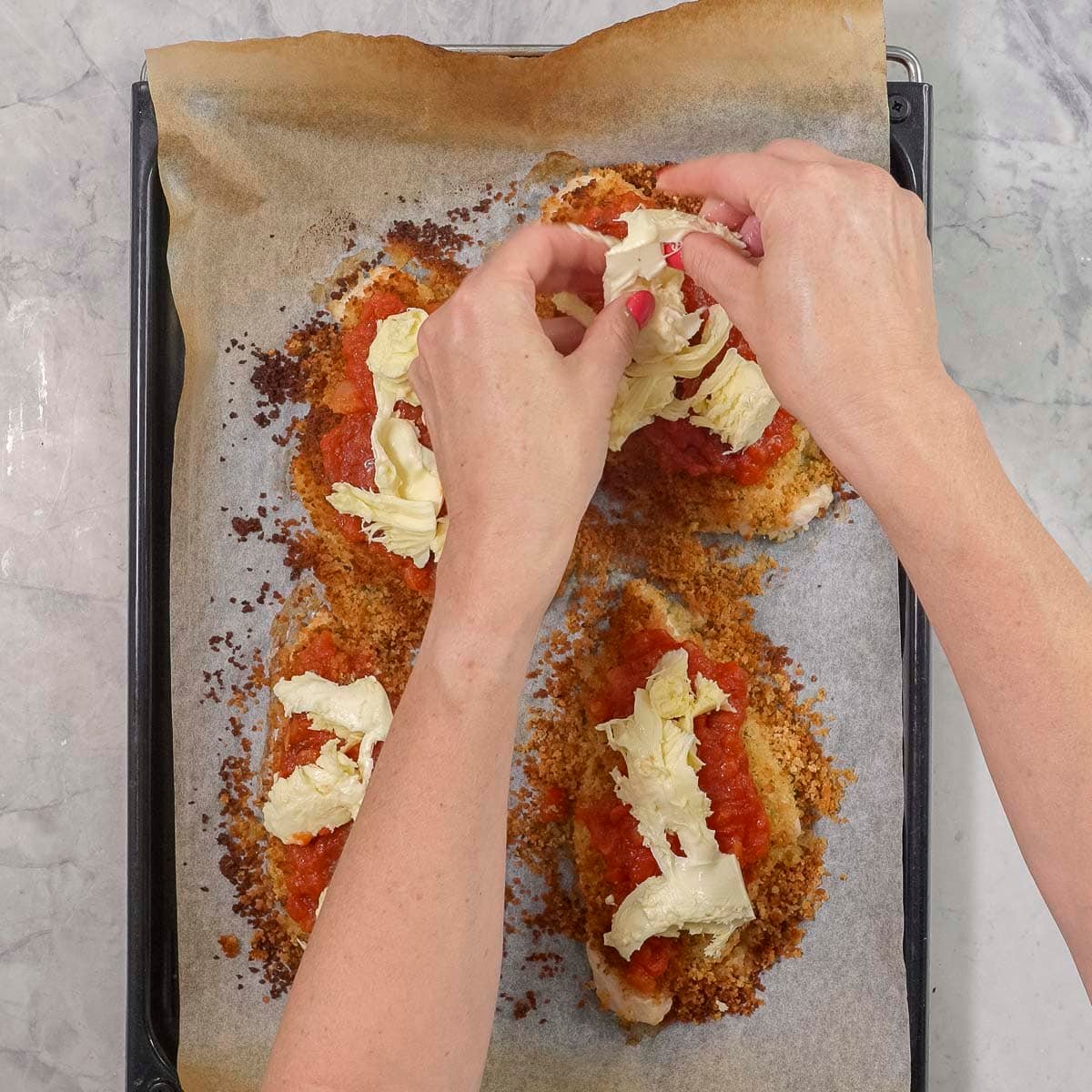 Hands placing mozzarella on top of four crispy parmesan chicken breasts.
