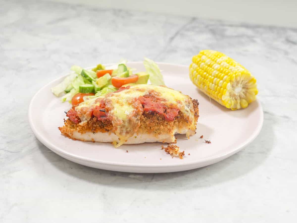 Plate on benchtop with chicken parmesan breast, fresh salad and corn cob.