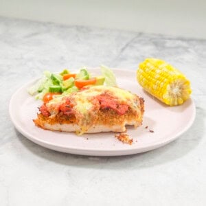 Plate on benchtop with chicken parmesan breast, fresh salad and corn cob.