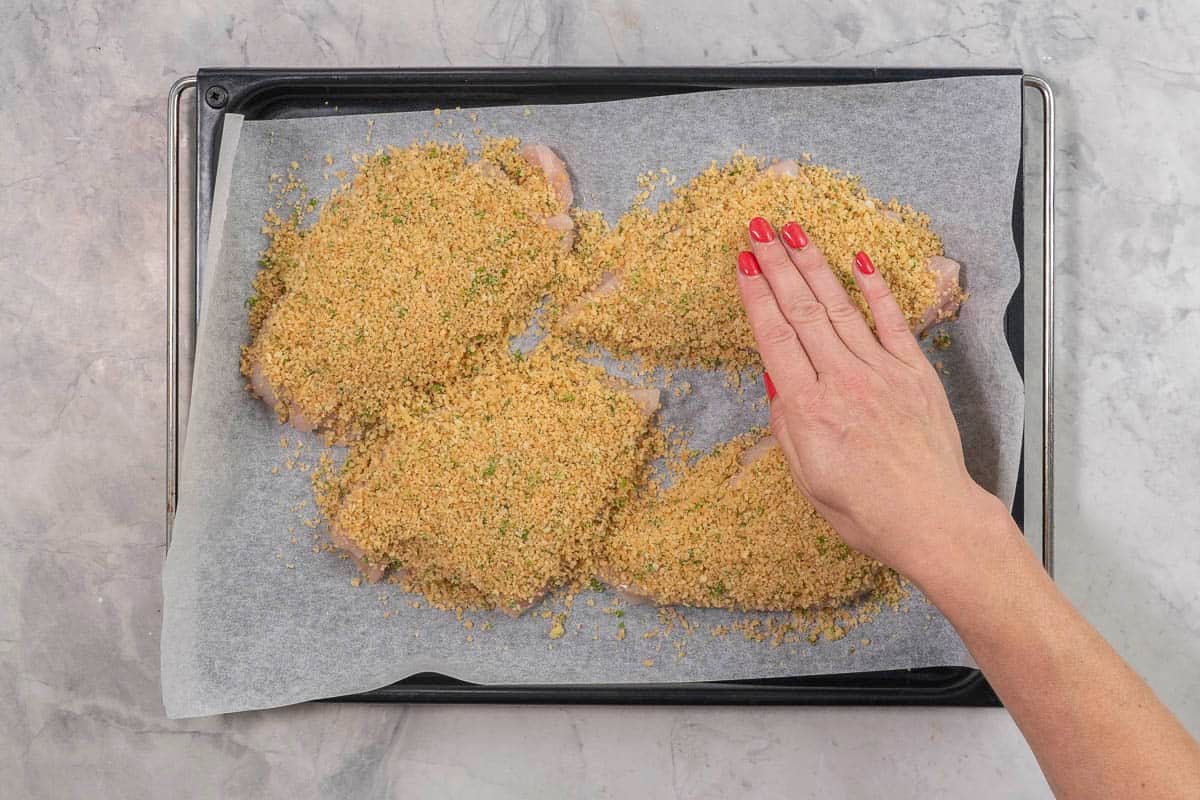 Hand patting down chicken parmesan coating onto four large chicken breasts on lined baking tray.