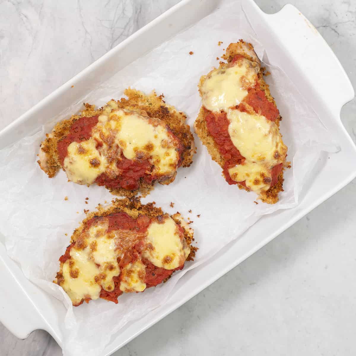 Three chicken breasts in a lined baking dish with paramasan coating, tomato and melted cheese on top.