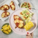Hand holding a plate with chicken parmesan breast, fresh salad and corn cob.