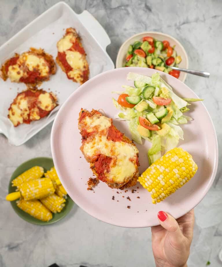 Hand holding a plate with chicken parmesan breast, fresh salad and corn cob.