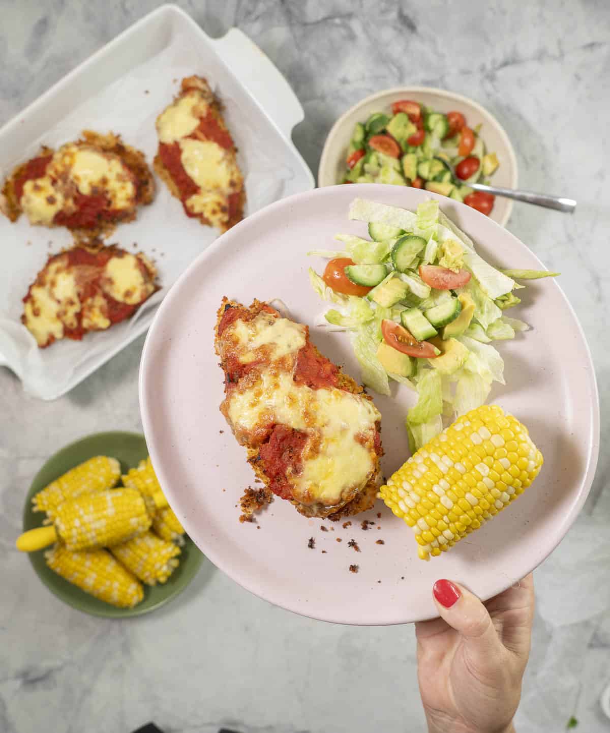 Hand holding a plate with chicken parmesan breast, fresh salad and corn cob.
