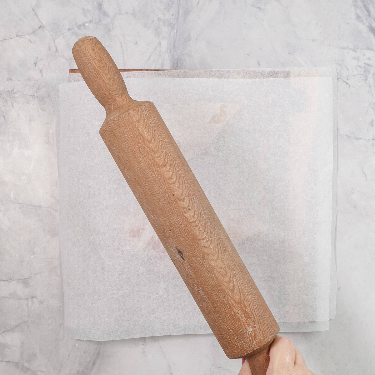 Hand holding a wooden rolling pin to flatten chicken breasts in between baking paper.