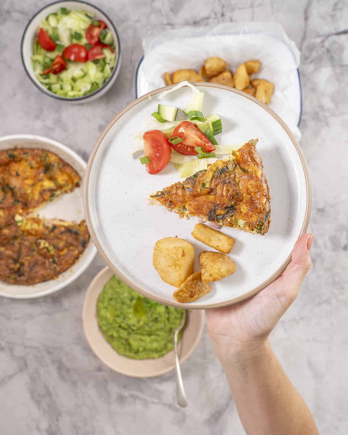 Hand holding a dinner plate with quiche fresh salad and roast potato.