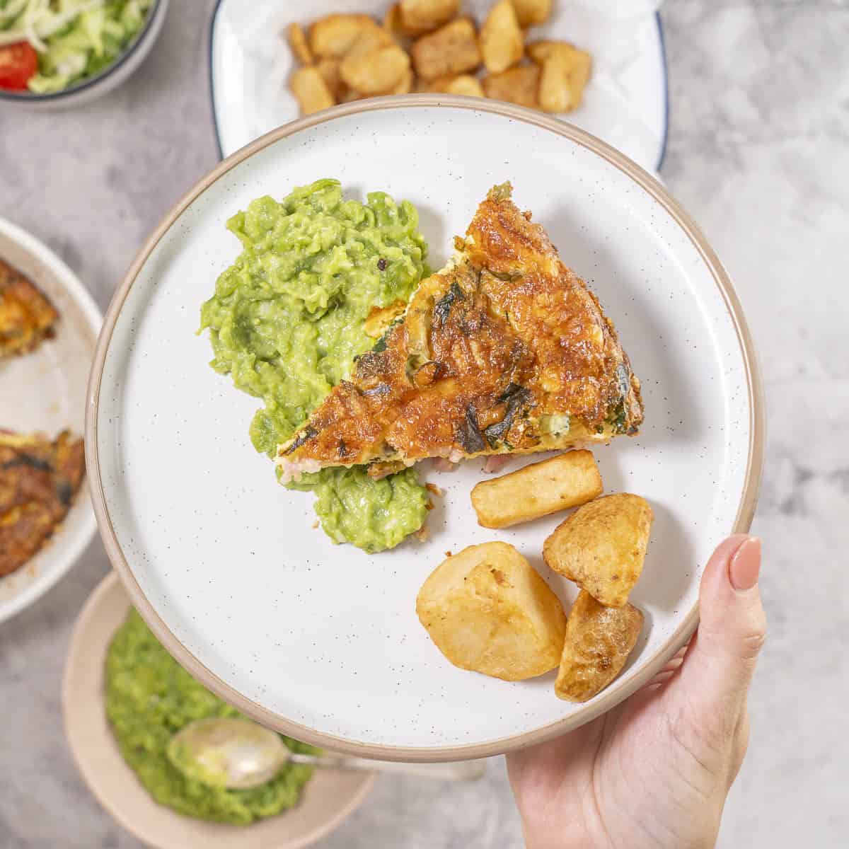 Hand holding a dinner plate with quiche, mashed peas and roast potato.