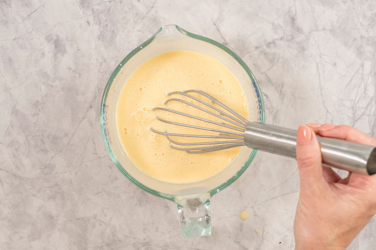 Hand holding a whisk in a glass measuring jug with corn flour, cream, eggs, garlic powder, mustard and salt inside whisked together.