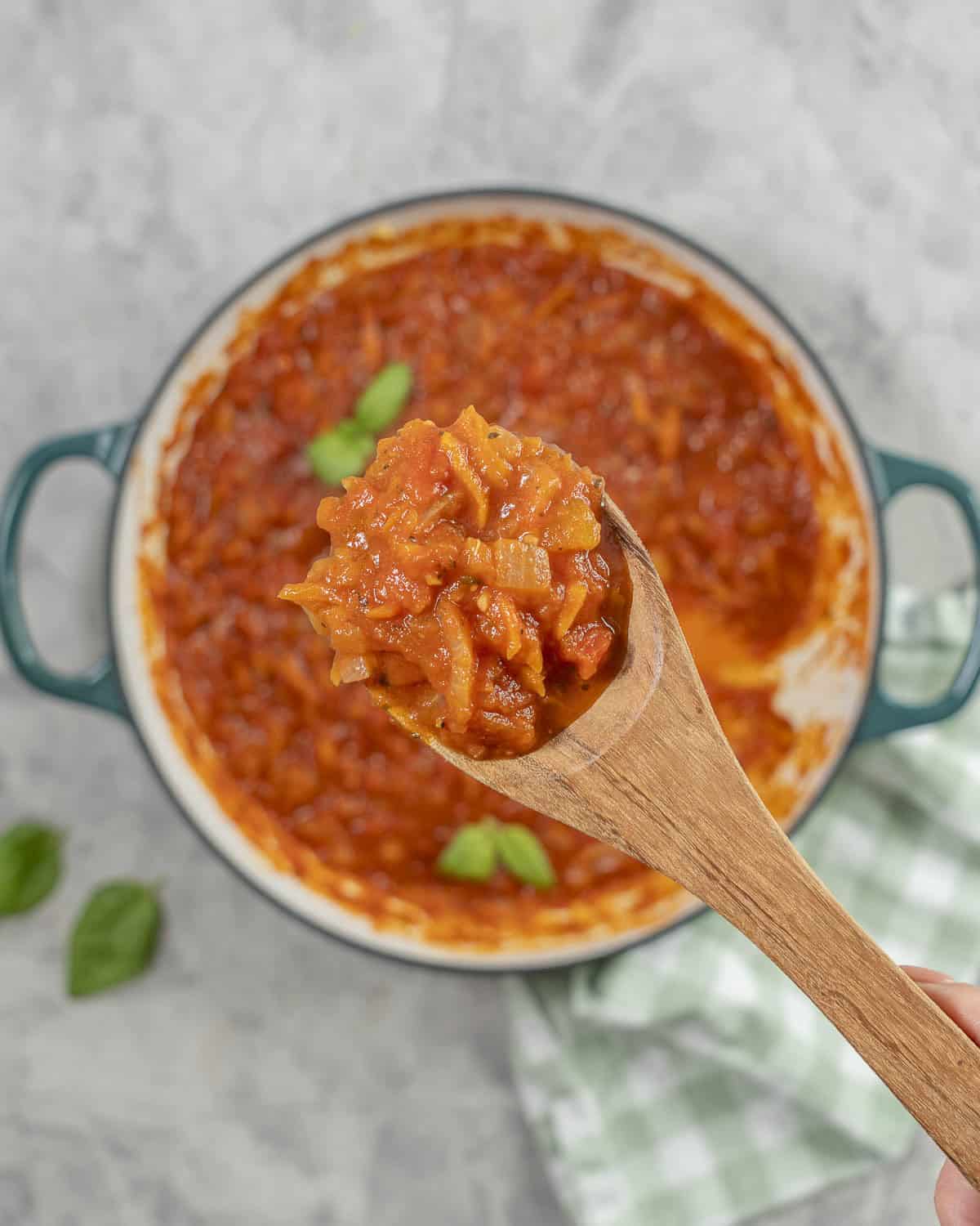 Hand holding a wooden spoon of marinara sauce over a pan of tomato sauce.