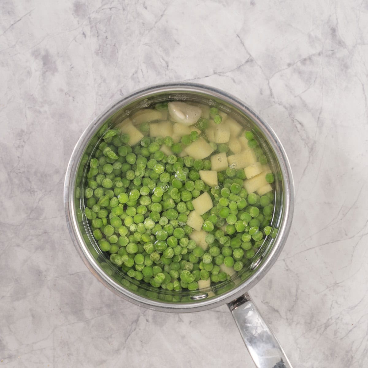 Frozen peas, chopped potato and garlic in saucepan with water on benchtop.