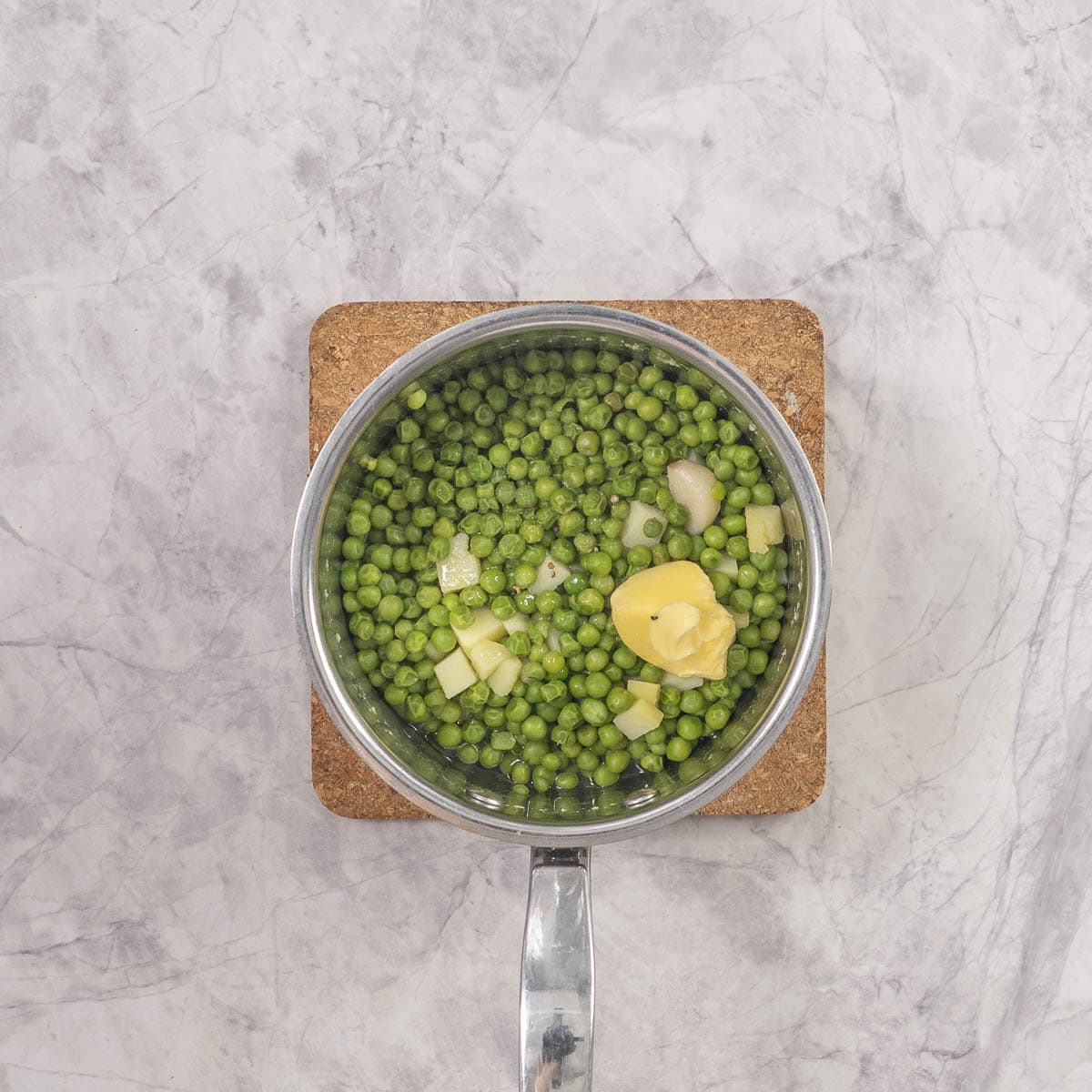 Saucepan placed on a cork mat on benchtop with Cooked peas, potato and garlic with butter and olive oil.
