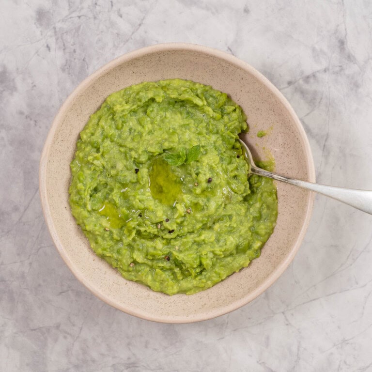 Mashed peas in a bowl with spoon garnished with piece of parsley and cracked pepper.