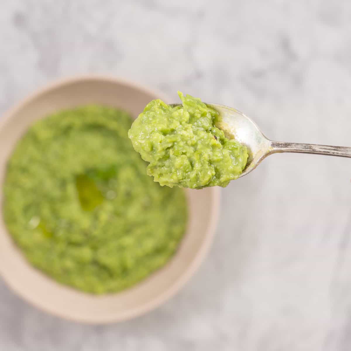 Mashed peas on a dessert spoon held up to camera with bowl of mashed peas in background on benchtop.