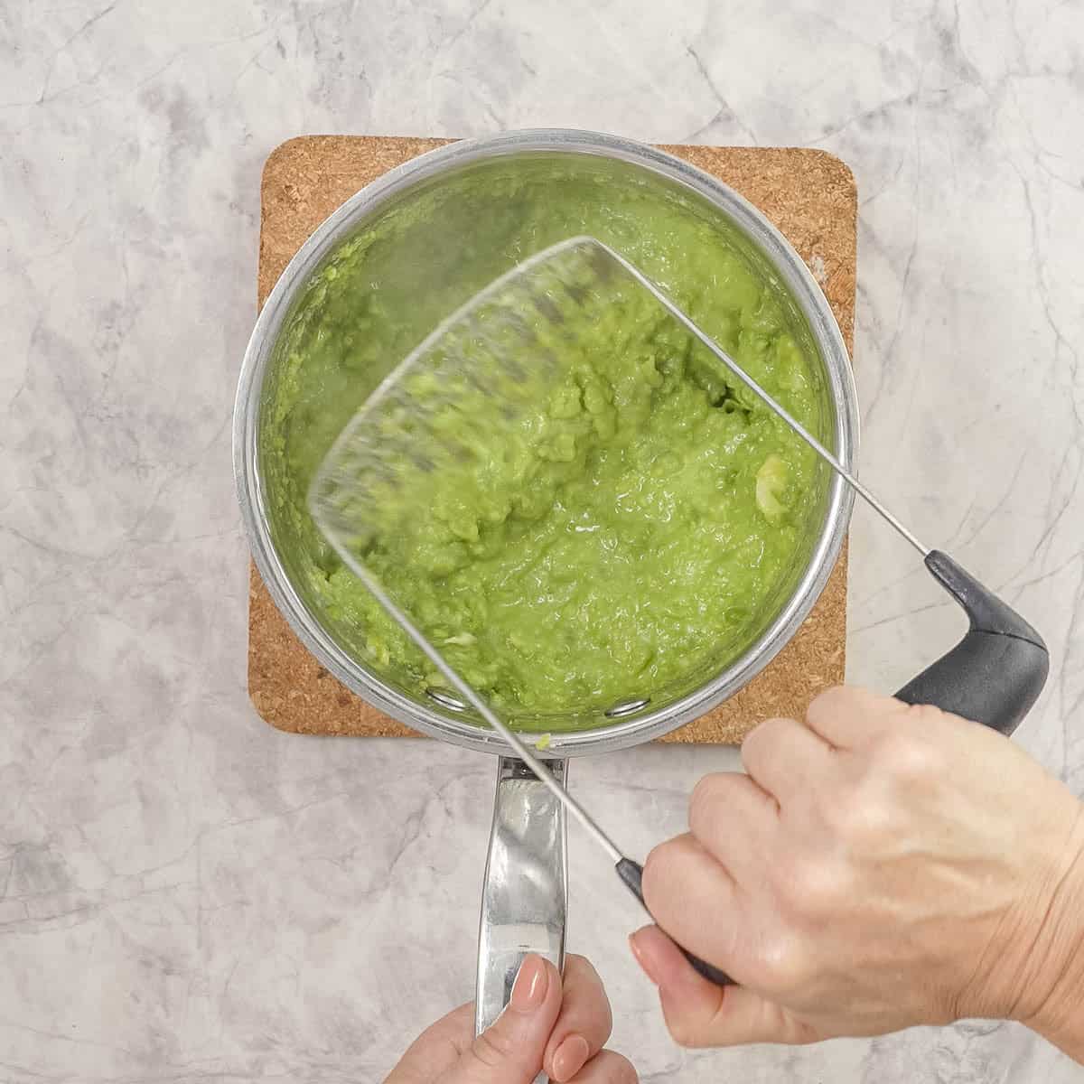 Hand holding a masher, mashing peas in saucepan.