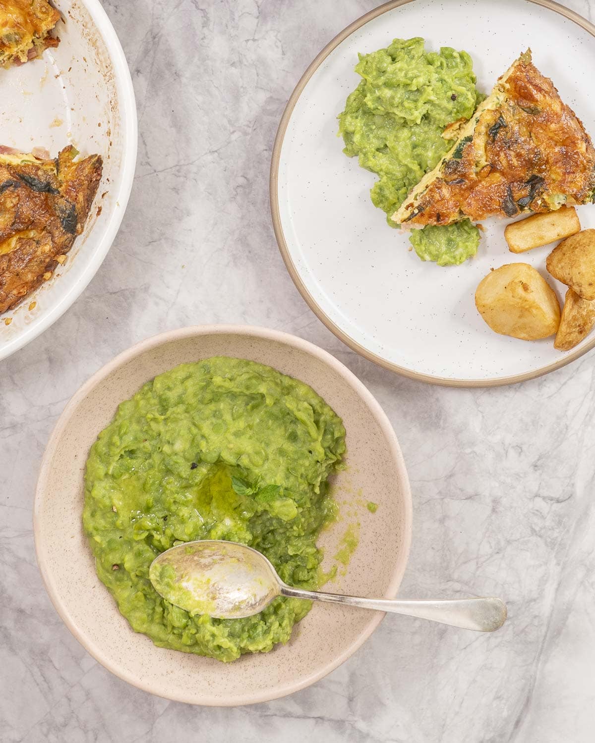 Mashed peas in a bowl with spoon, dinner plate with quiche, roast potato and mashed peas.