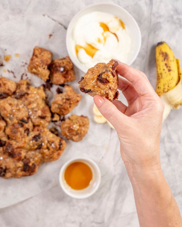 Womans hand holding a piece of monkey bread up to camera with monkey bread, yogurt and banana in background on bench top.