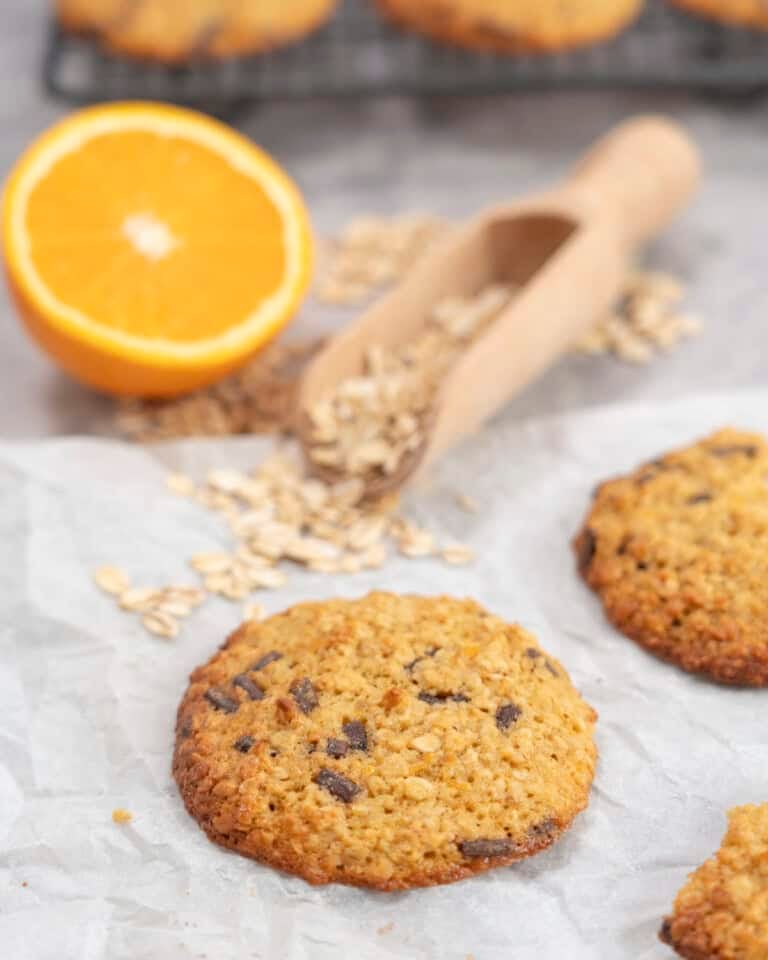 Orange oatmeal cookie with golden edges, wooden scoop of oats and halved orange on benchtop.