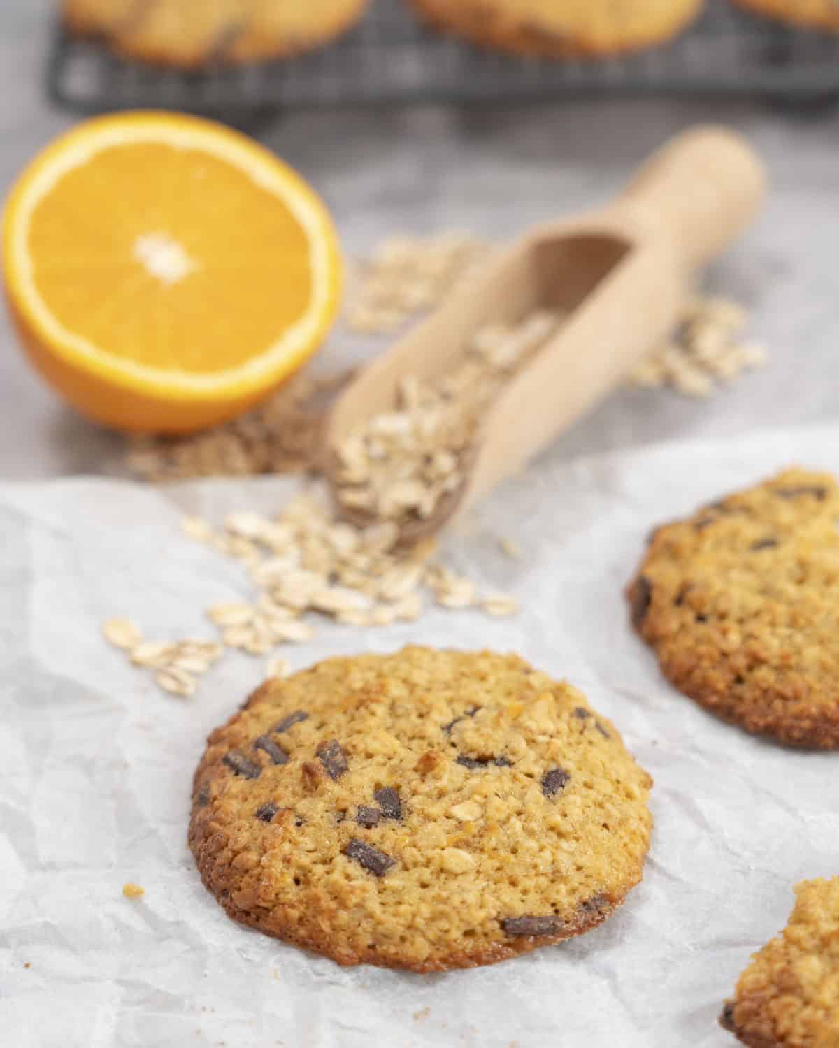 Orange oatmeal cookie with golden edges, wooden scoop of oats and halved orange on benchtop.