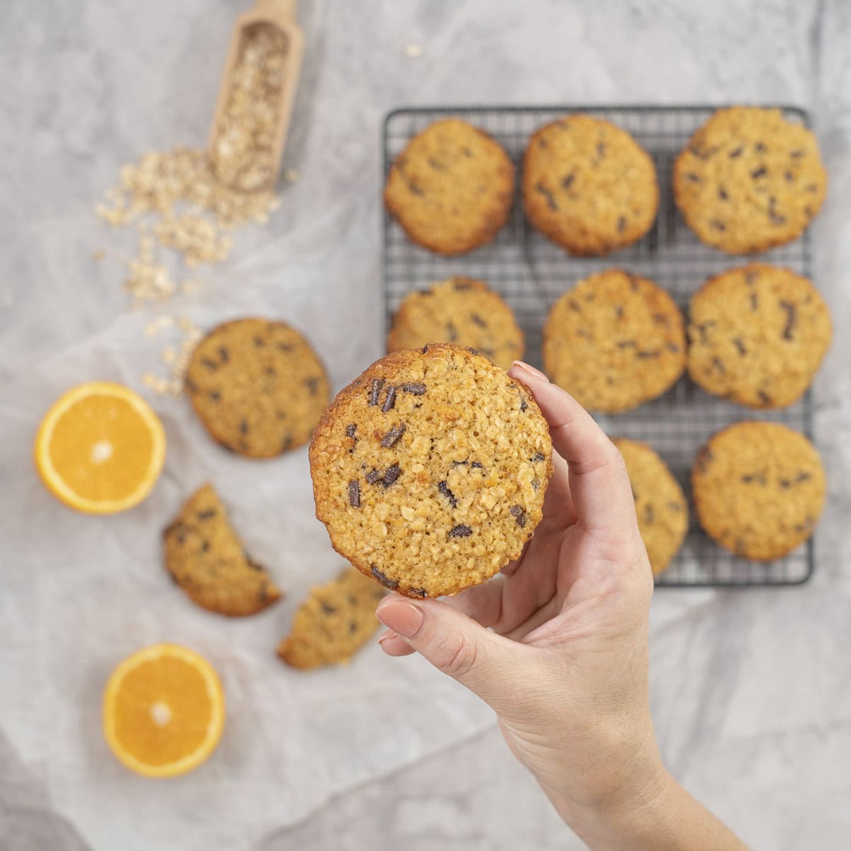 Hand holding a cookie up to camera, cooling tray with nine cookies below, orange cut in half and cookie broken in half laid on piece of baking paper.