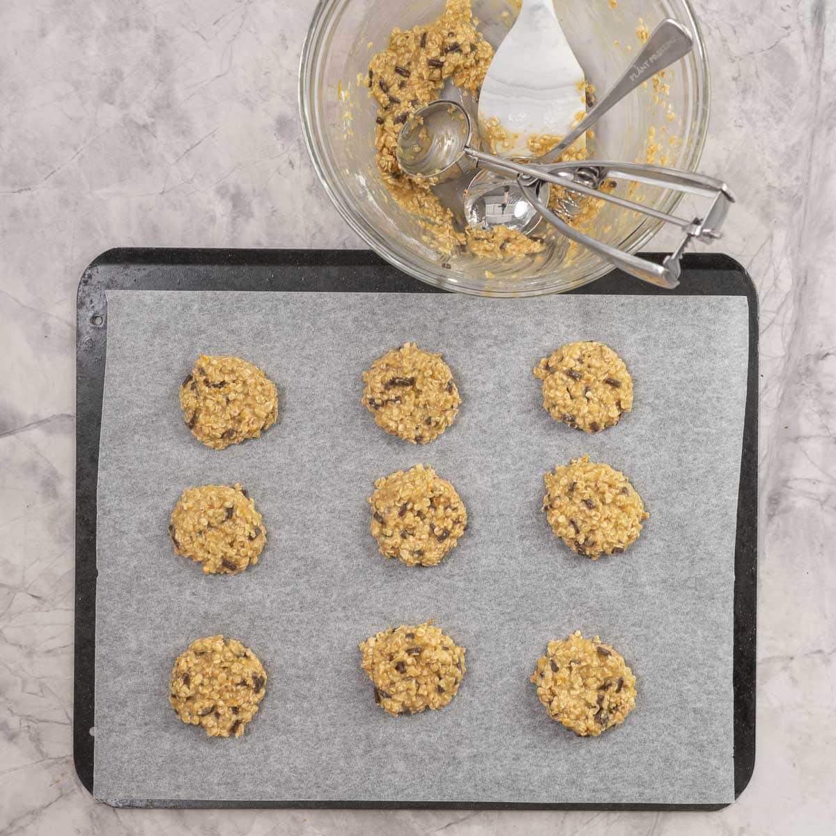 Nine scoops of cookie batter on lined baking tray on benchtop, empty bowl of batter and cookie scoops.