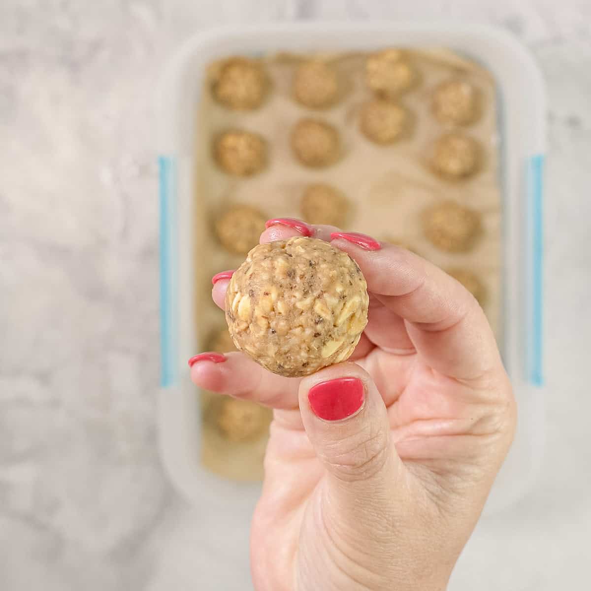 Hand holding up a peanut butter oat ball with container blurred in background with more inside.