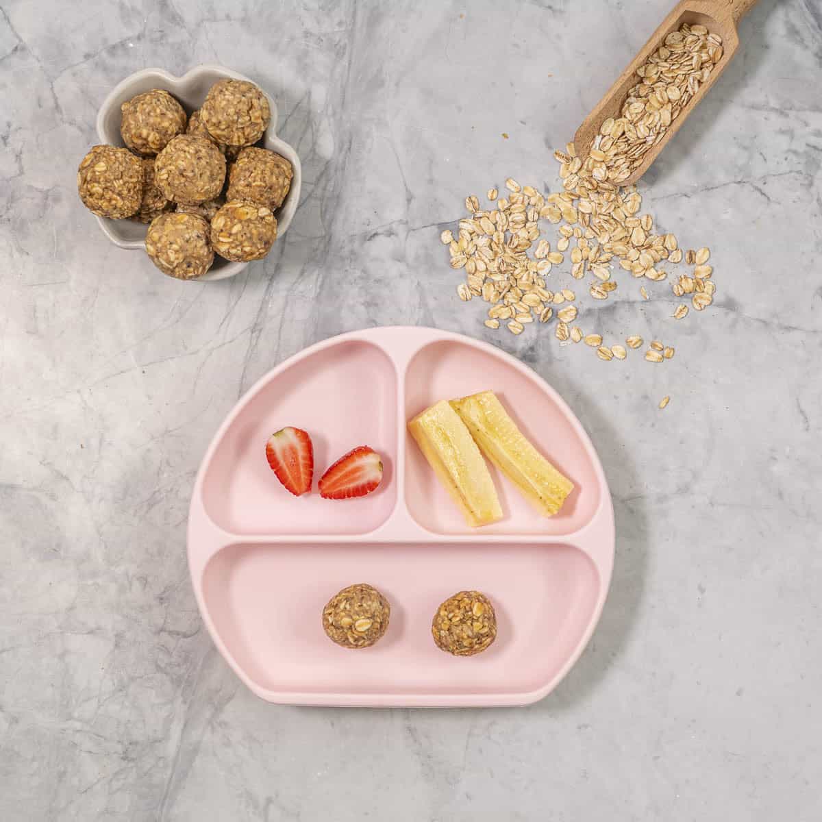 Pink Silicone baby plate on benchtop with half a strawberry, halved banana slices and two peanut butter oat balls. Ramekin with oat balls inside, wooden scoop with oats inside on bench top.