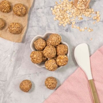 Peanut butter oat balls on benchtop with small ramekin with more balls inside, wooden scoop with oats on bench top and spatula.