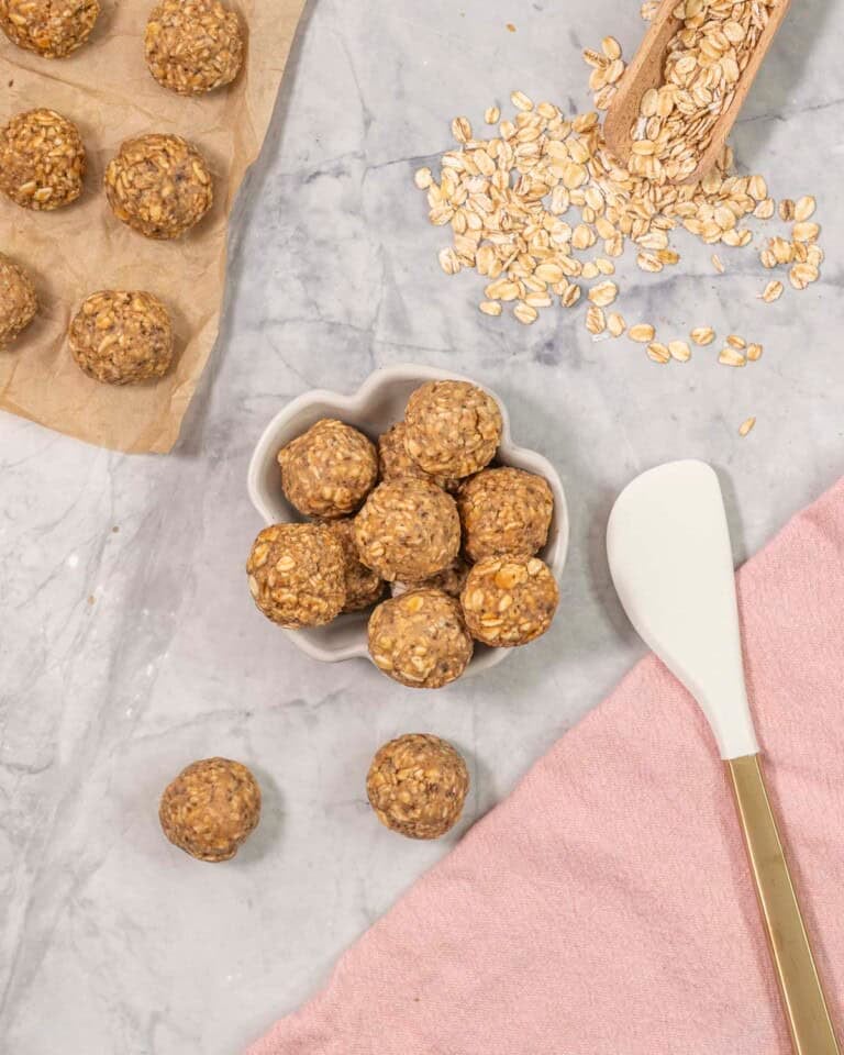 Peanut butter oat balls on benchtop with small ramekin with more balls inside, wooden scoop with oats on bench top and spatula.
