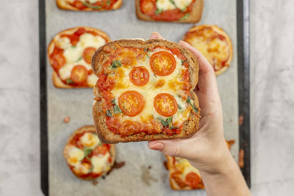A slice of pizza toast topped with golden cheese, tomato slices and shredded spinach being held above a tray of more cooked pizzas.