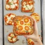A slice of pizza toast topped with golden cheese, tomato slices and shredded spinach being held above a tray of more cooked pizzas.