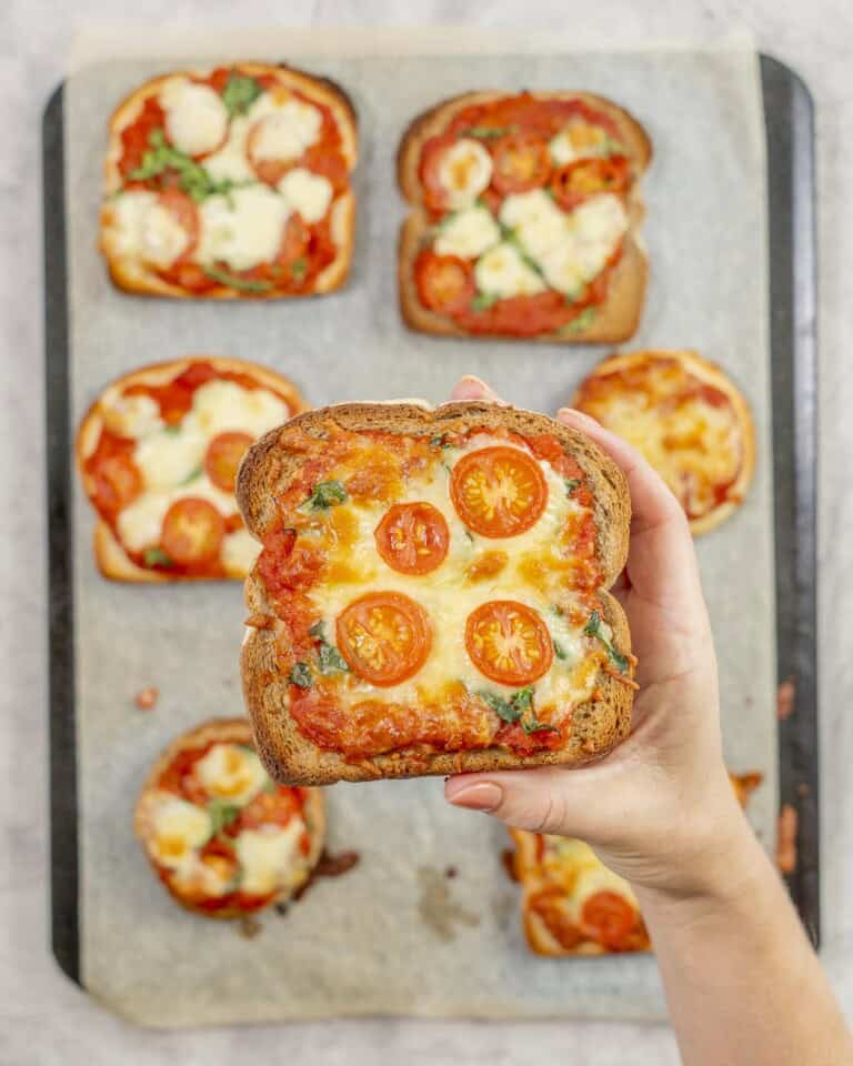 A slice of pizza toast topped with golden cheese, tomato slices and shredded spinach being held above a tray of more cooked pizzas.