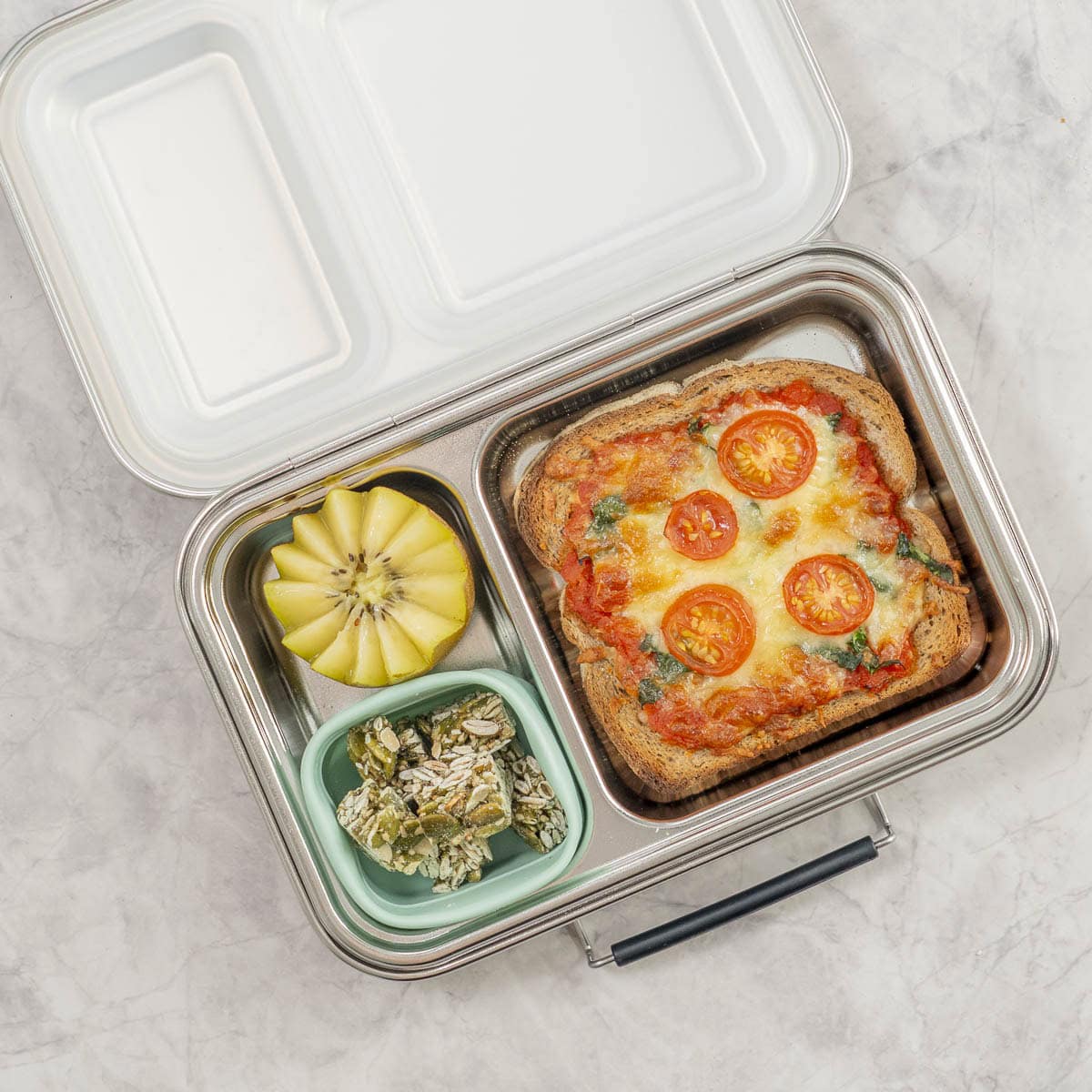 A pizza toast packed into a stainless lunch box with a kiwi and seed snacks.
