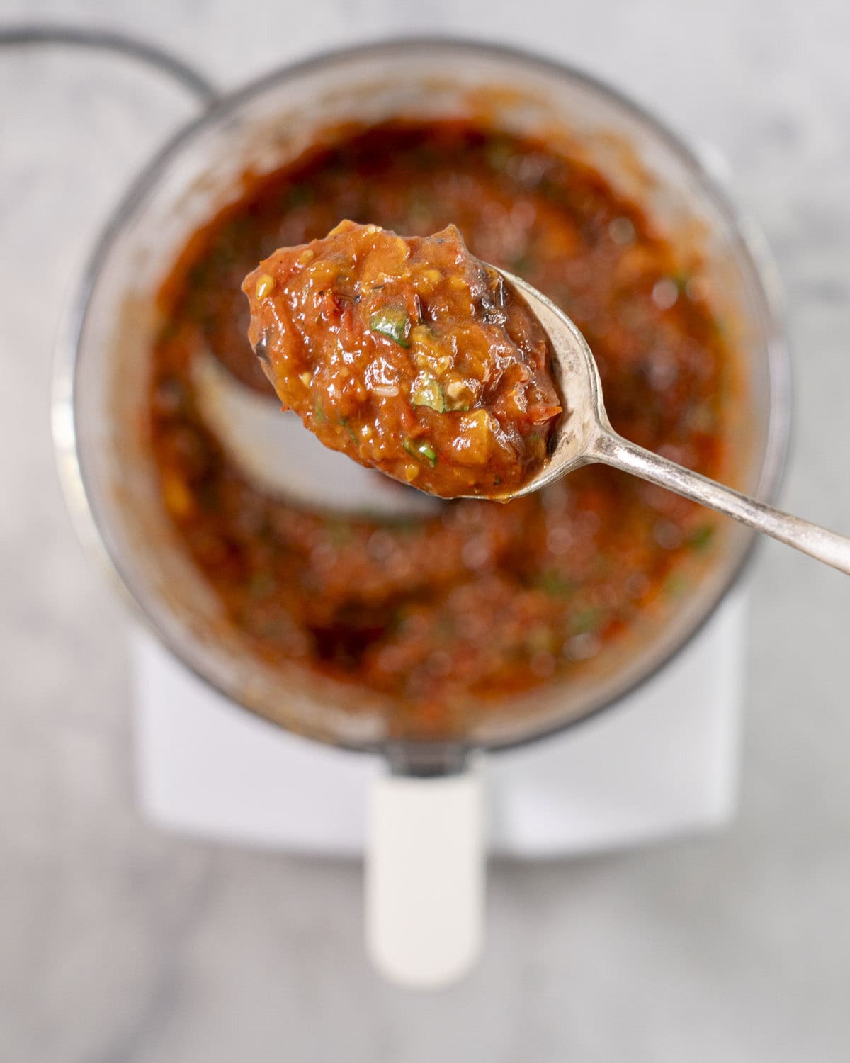 Spoon with roasted tomato sauce held above food processor with toasted tomato sauce inside.